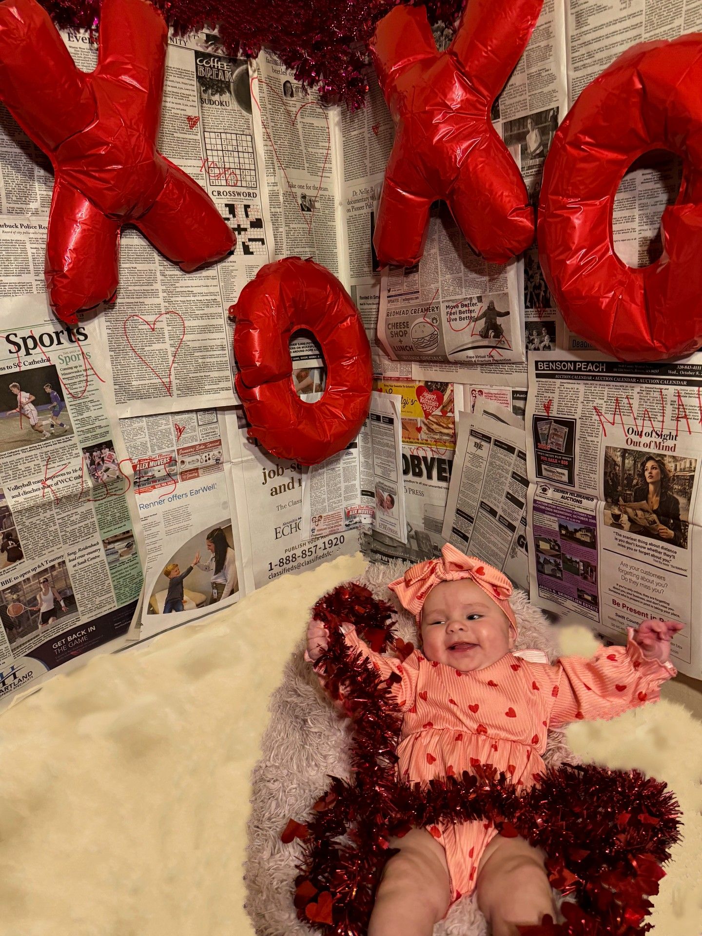 Infant in pink heart outfit on white rug with red tinsel and XOXO balloon Valentine's Day decor.