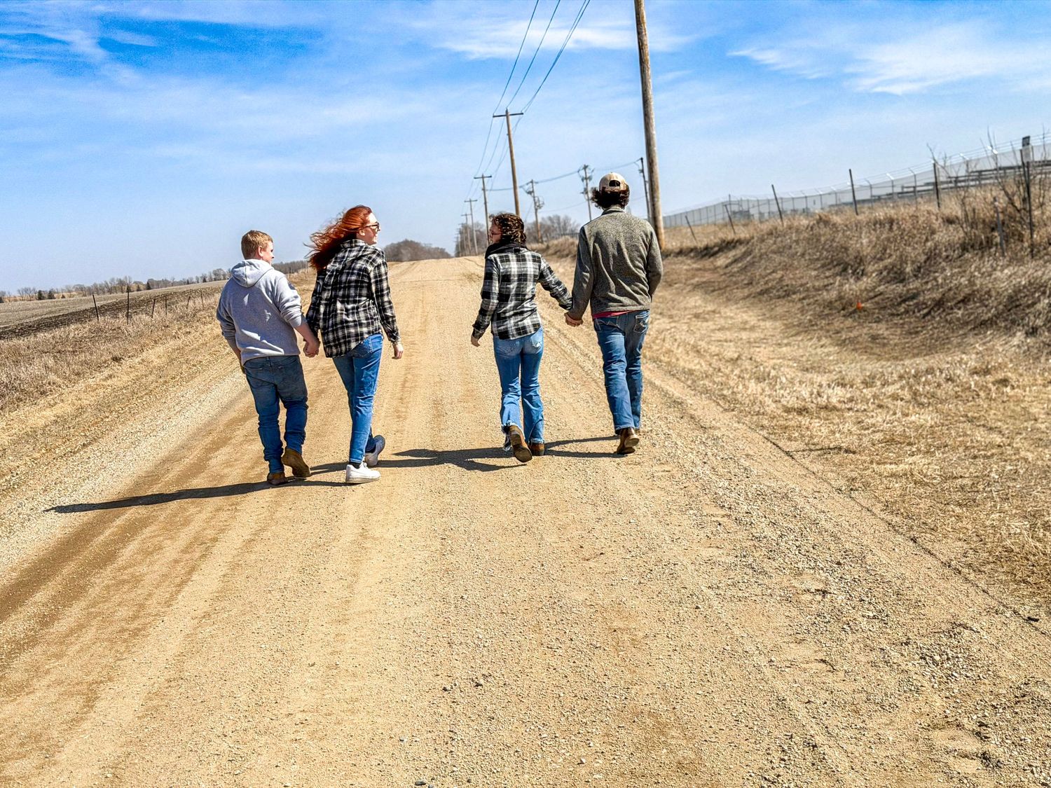 Four people walking together holding hands down a wide open rural dirt road under blue sky.