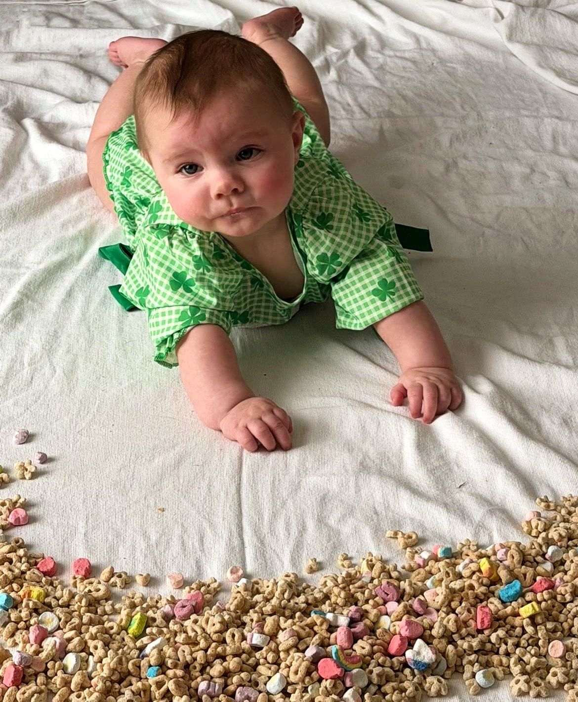 Baby in green shamrock romper doing tummy time near Lucky Charms cereal.