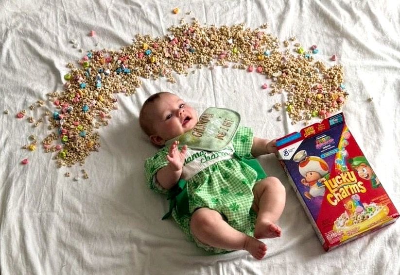 Smiling baby in green outfit laying beside Lucky Charms cereal box.
