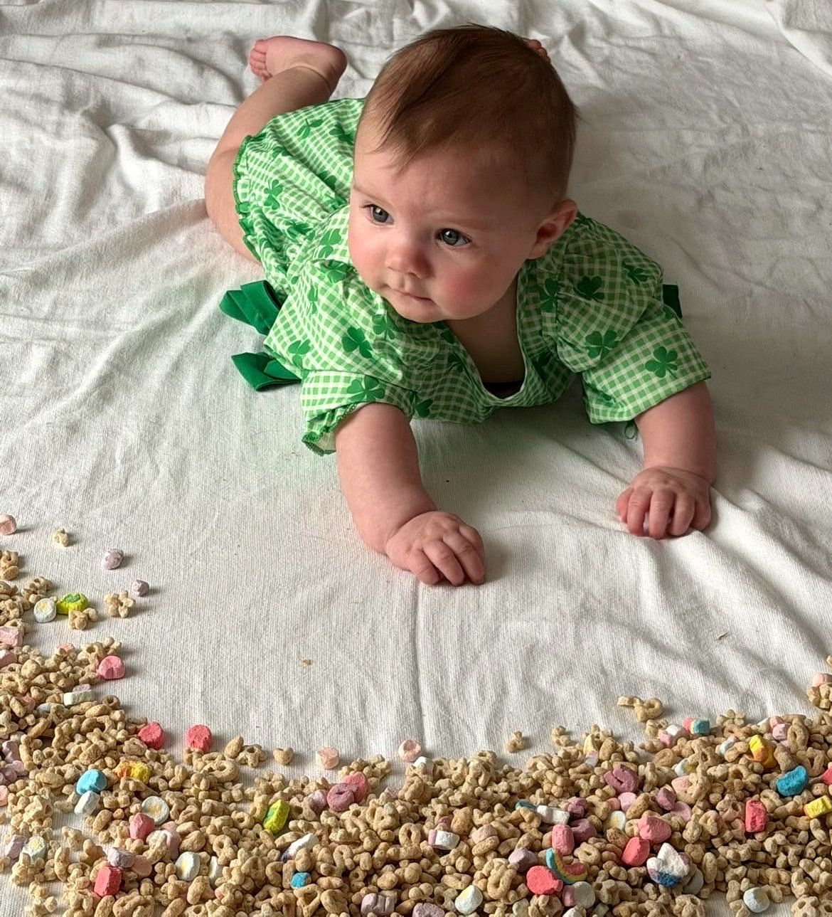 Baby in green gingham romper doing tummy time near Lucky Charms cereal.