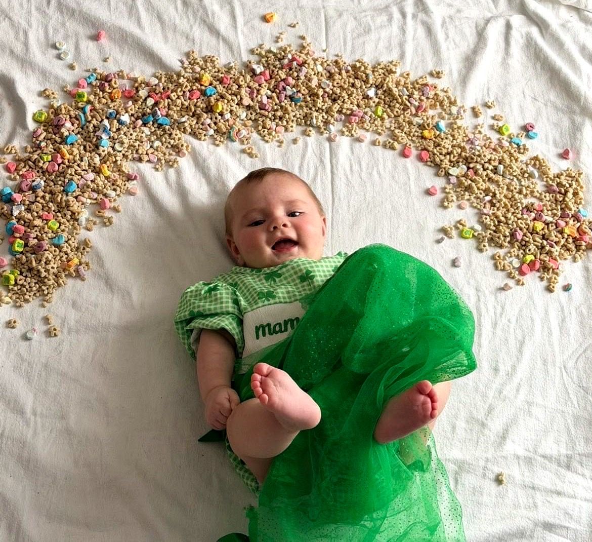Happy baby in green gingham outfit surrounded by Lucky Charms cereal rainbow.