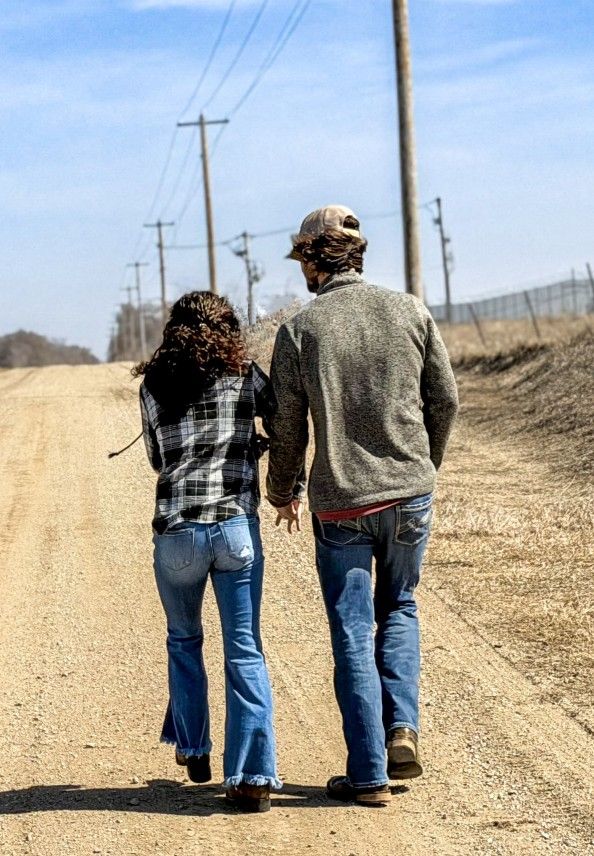 Couple walking hand in hand down rural dirt road lined with utility poles on a sunny day.