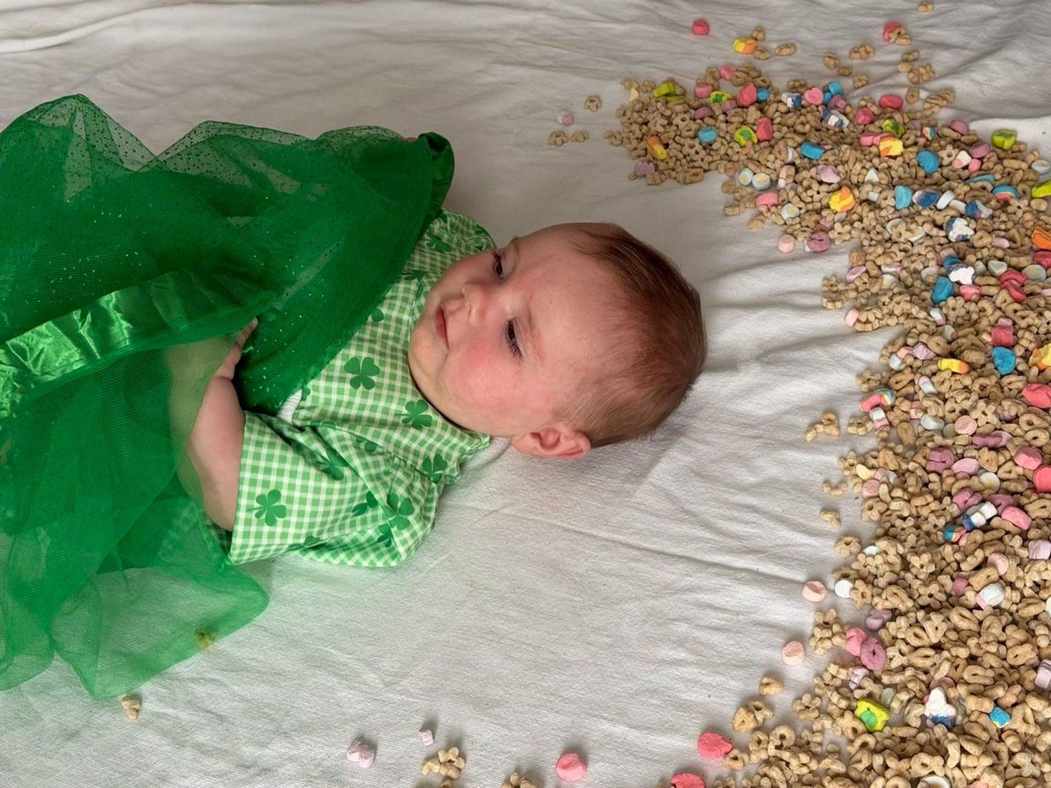 Baby in green gingham outfit laying beside colorful Lucky Charms cereal pile.