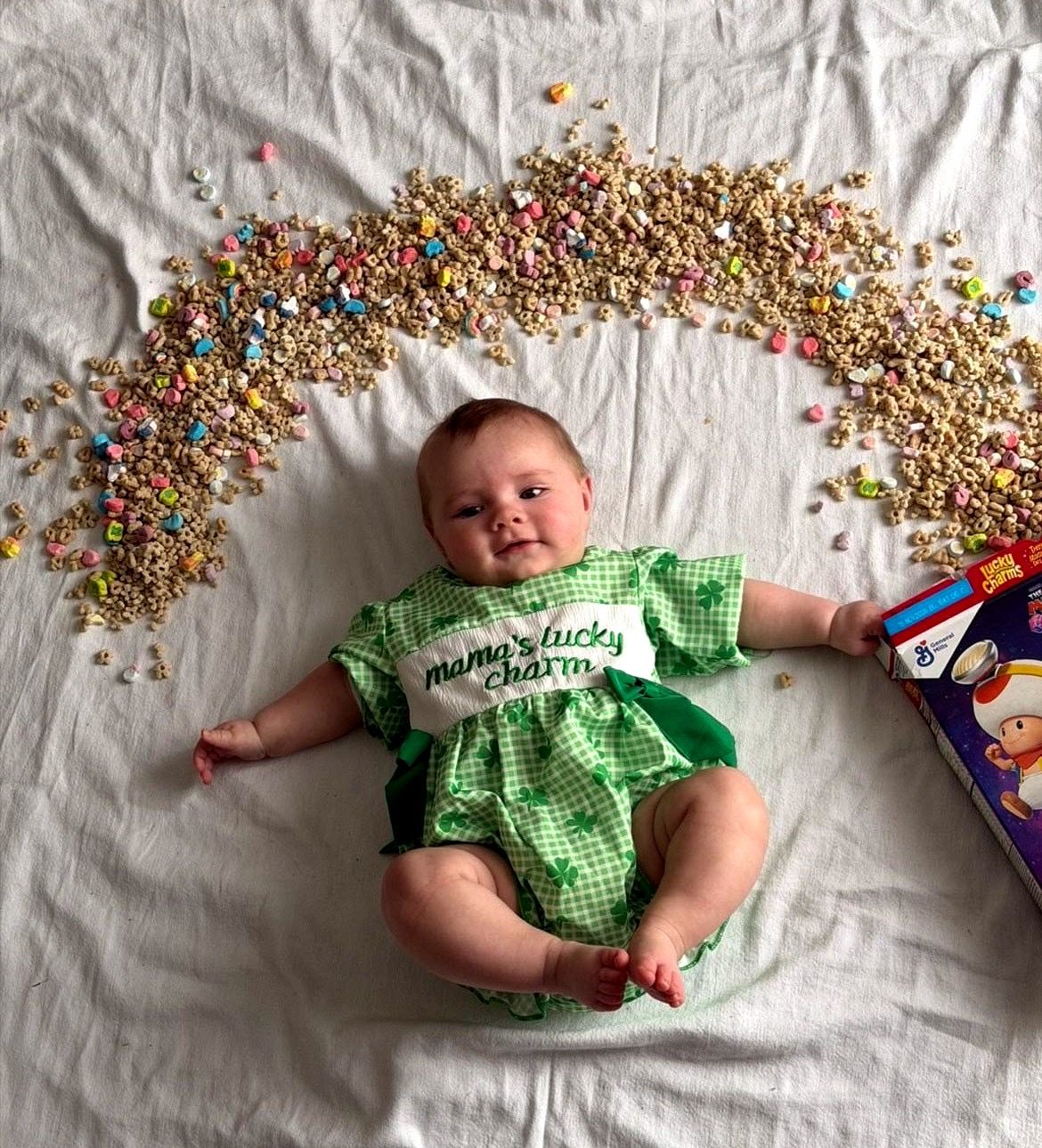 Smiling baby in green gingham 'mama's lucky charm' outfit surrounded by Lucky Charms cereal arc.