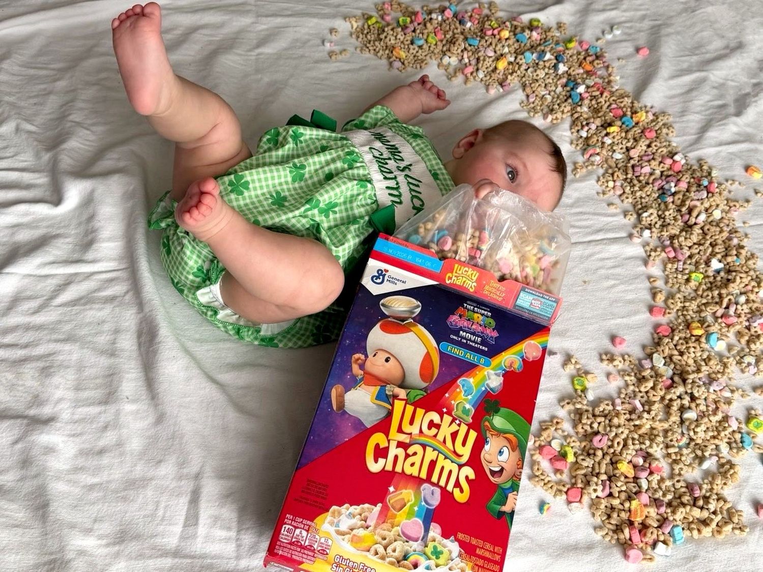 Baby in green outfit lying next to Lucky Charms cereal box with colorful marshmallows scattered around.
