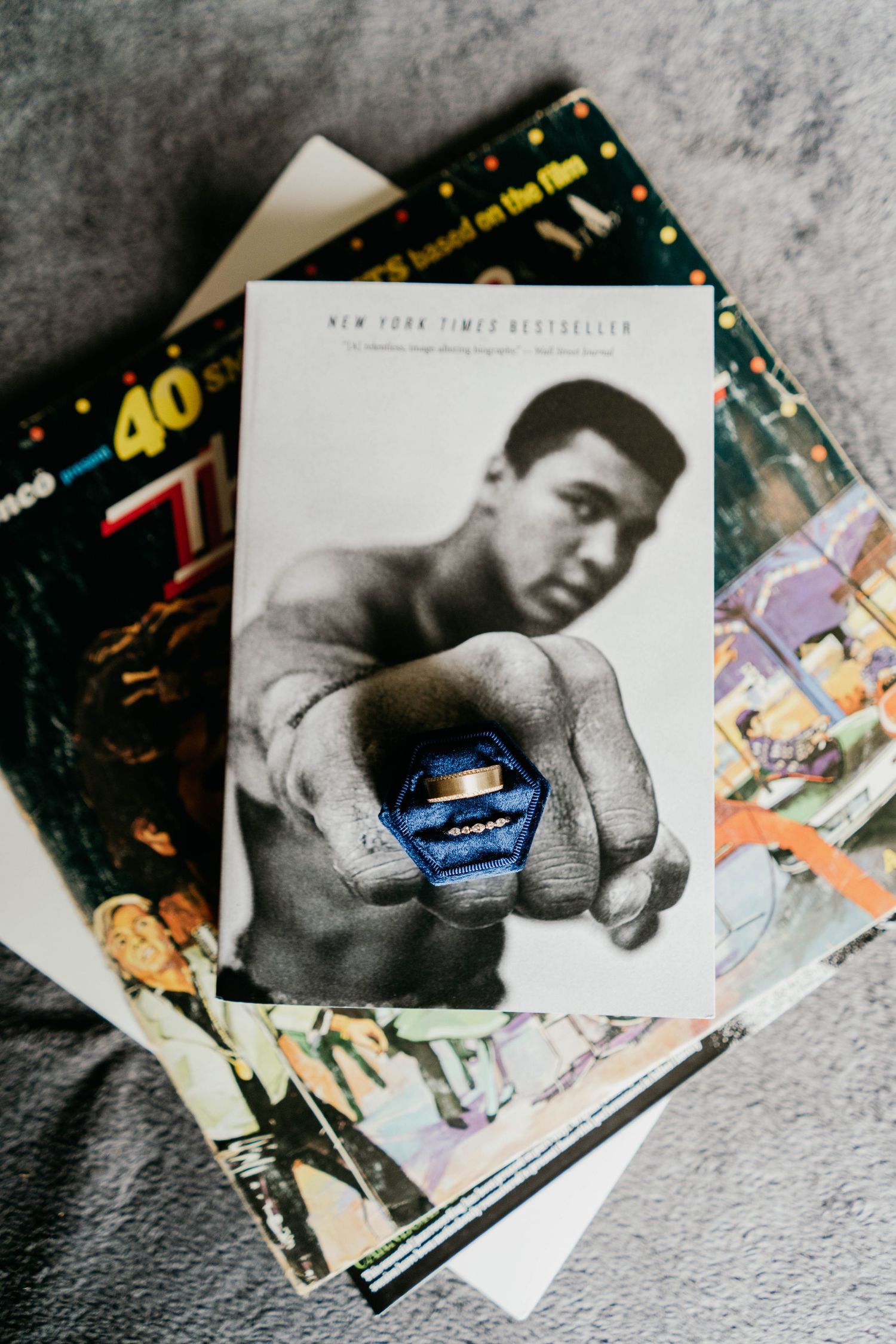 Detail shot of wedding ring bands on top of a Mohammad Ali biography book stacked on top of vintage records set in a blue velvet ringbox