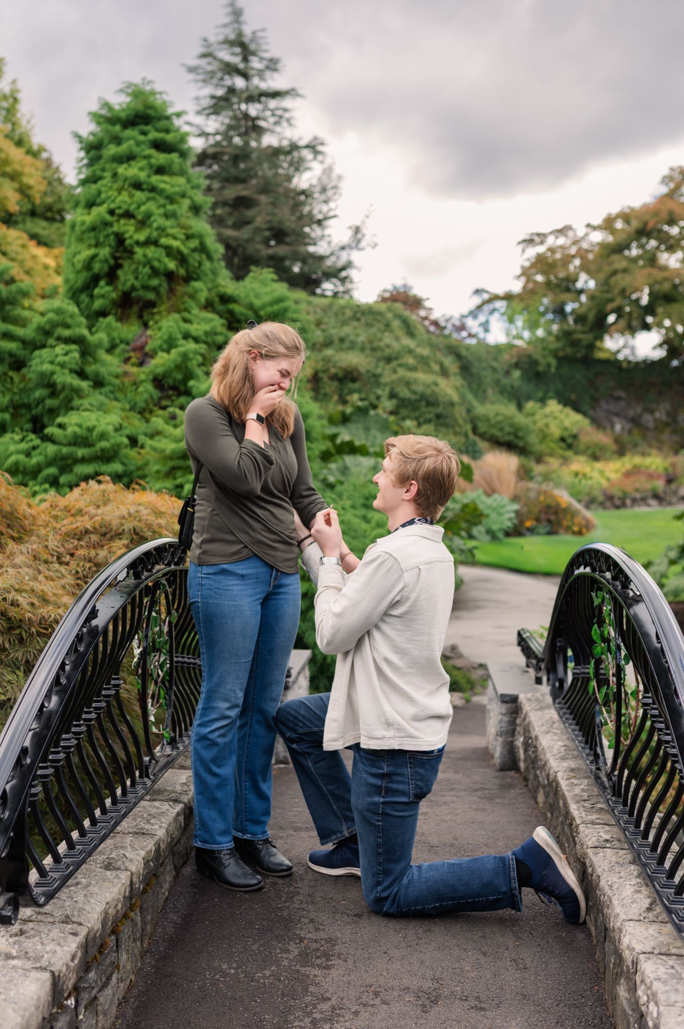 Person proposing on one knee to their partner on a scenic garden bridge, surrounded by lush green trees at Queen Elizabeth park in Vancouver