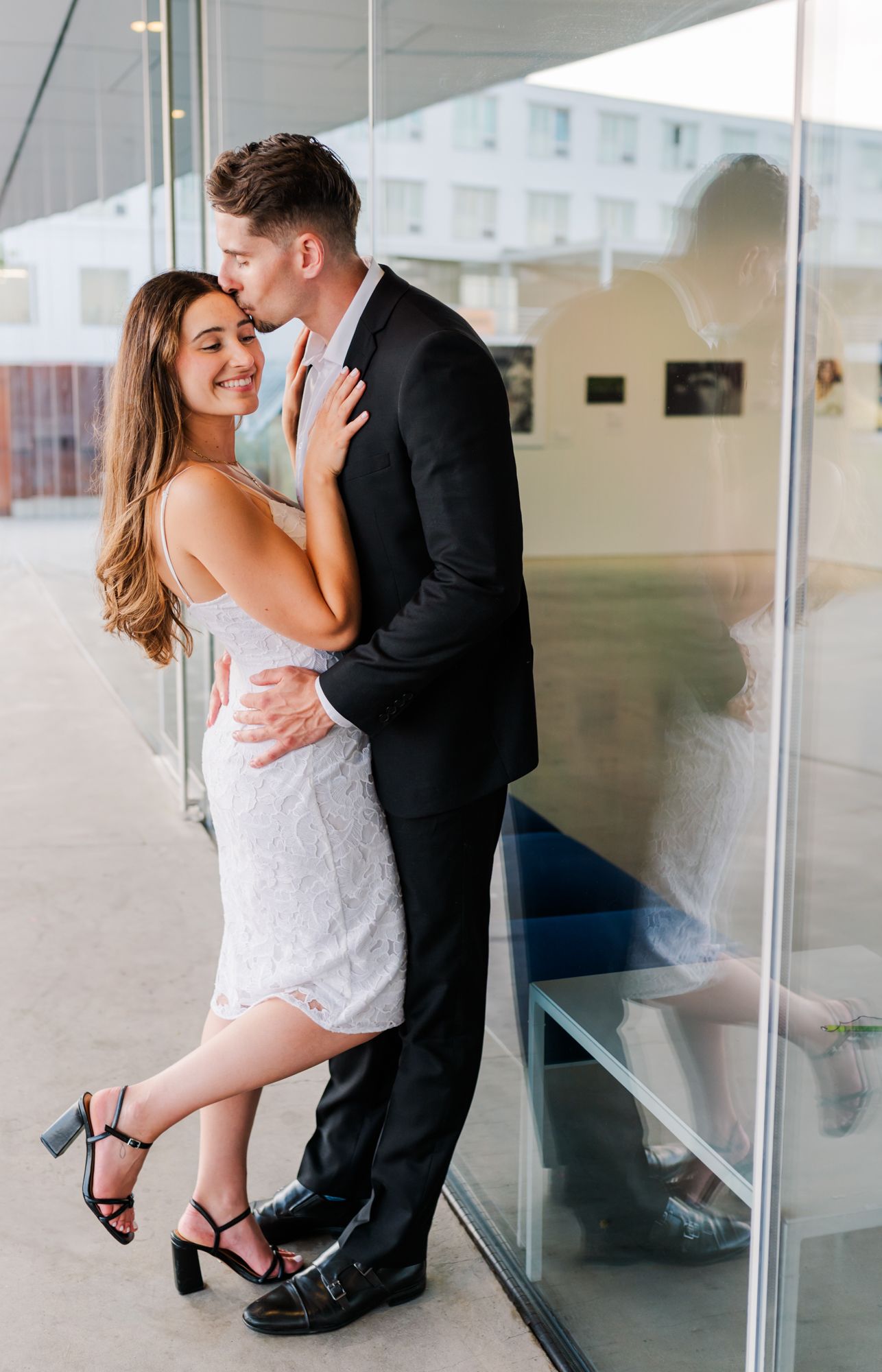 Couple embracing and kissing near glass wall, woman in white lace dress and heels, man in black suit in Vancouver, BC.