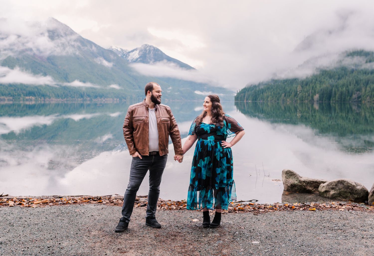 Couple holding hands by a misty mountain lake with stunning reflections, wearing casual and floral dress outdoors.