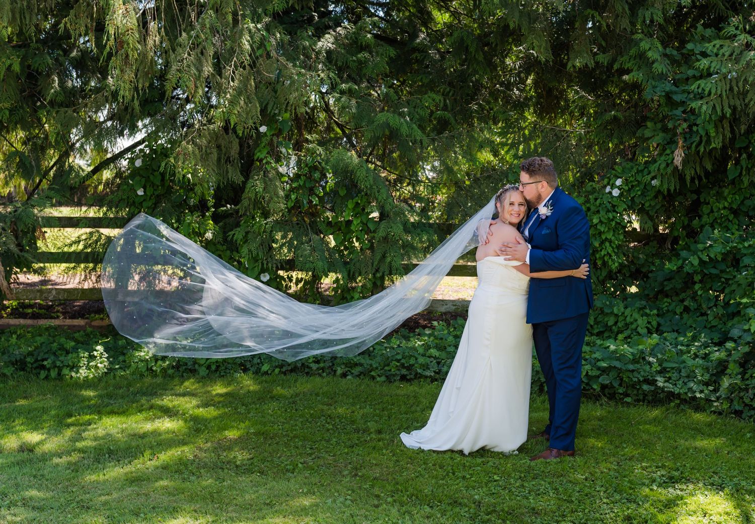 Bride and groom share a romantic moment outdoors, her long veil flowing in the breeze, surrounded by lush green trees Fraser Valley wedding.