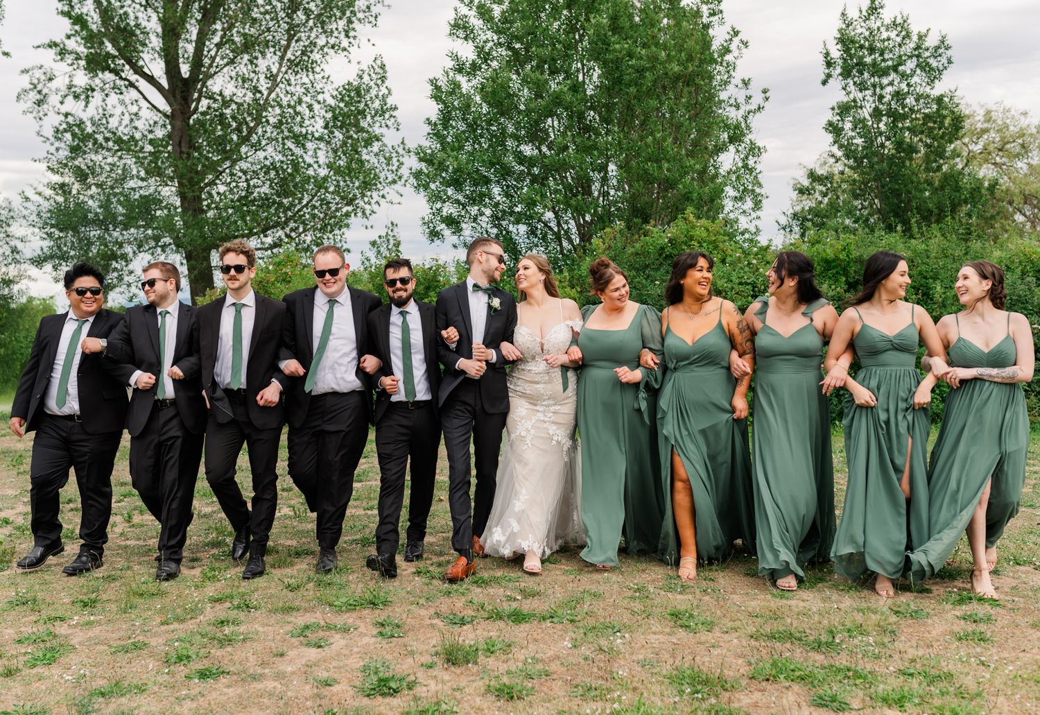Fraser Valley wedding party walks outdoors, groomsmen in black suits and bridesmaids in sage green dresses flanking the bride and groom.