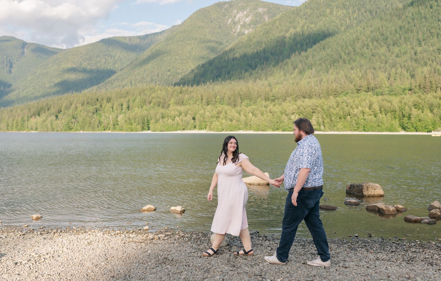 Couple holding hands walking along a rocky lakeside beach with lush green mountains in the background.