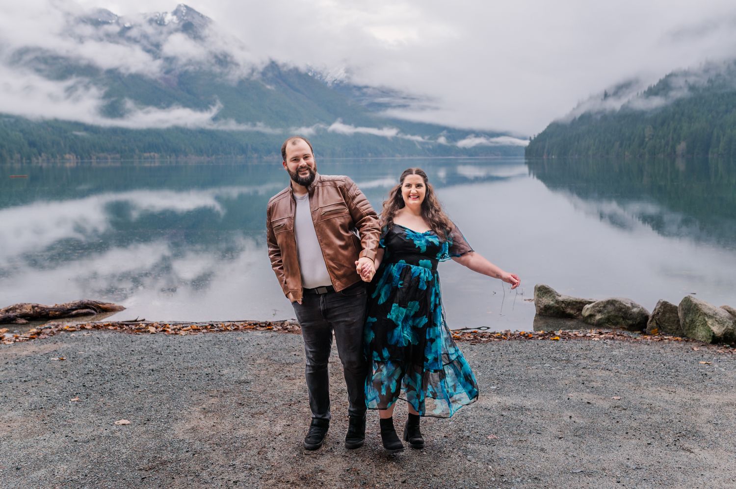Couple holding hands on their engagement smiling by a reflective mountain lake with misty peaks in the background at Chilliwack Lake.