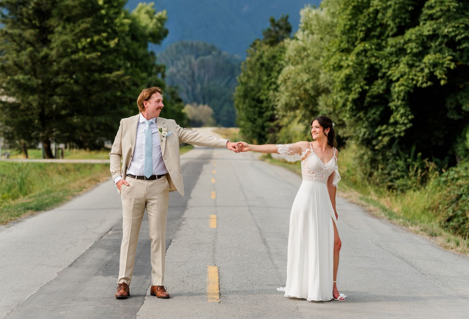 Bride and groom hold hands at arm's length on an empty road, surrounded by lush green trees on their Maple Ridge wedding day.
