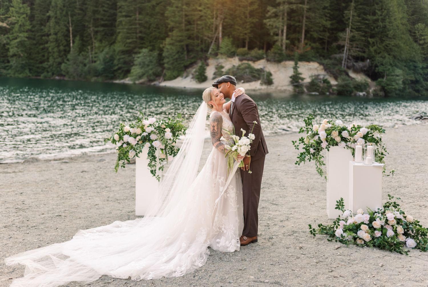 Bride and groom share a kiss at an outdoor lakeside Fraser Valley wedding ceremony with white floral arrangements and lush green trees.