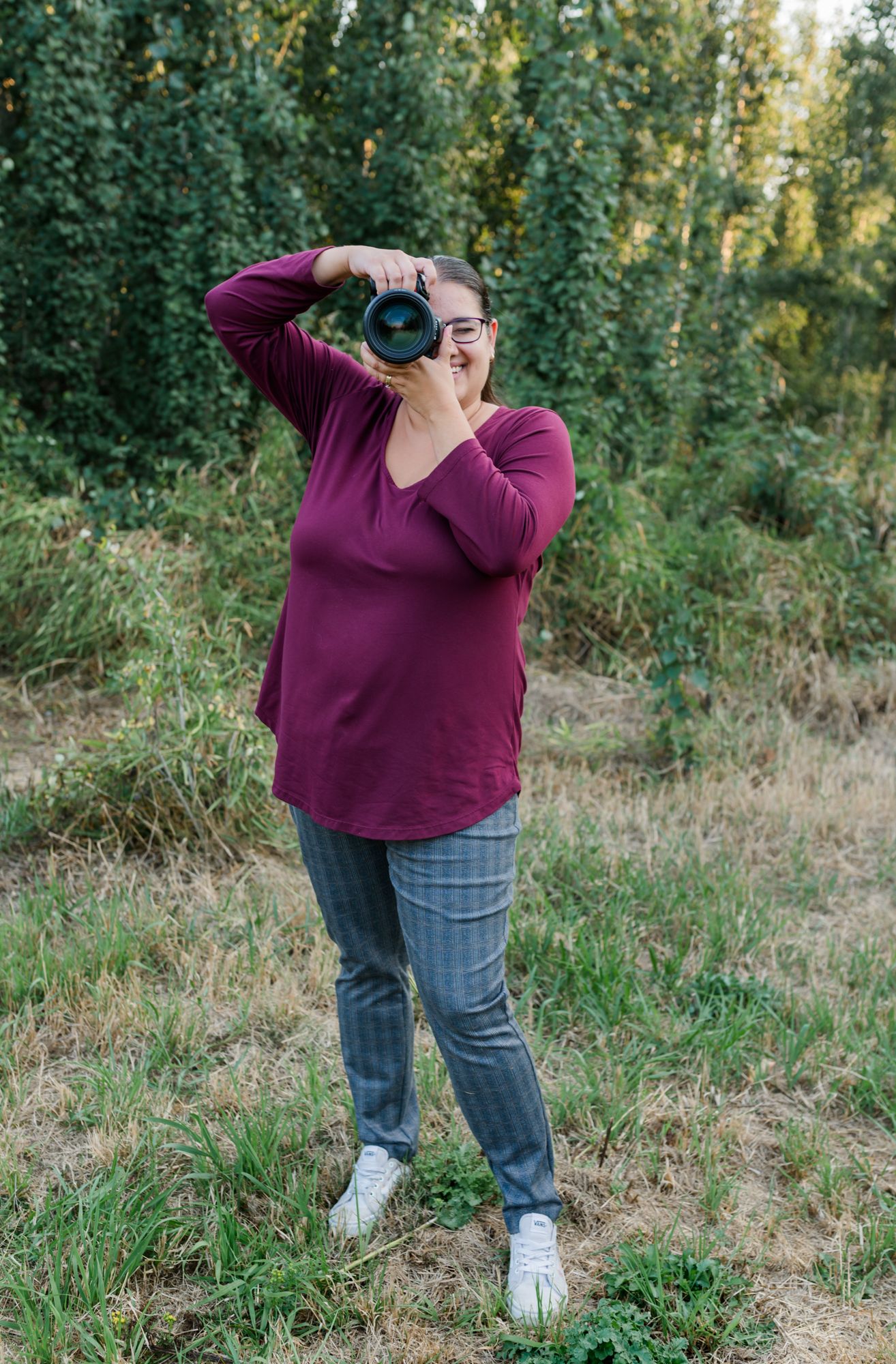 Fraser Valley photographer in burgundy top holds DSLR camera up to face while standing in green field outdoors.