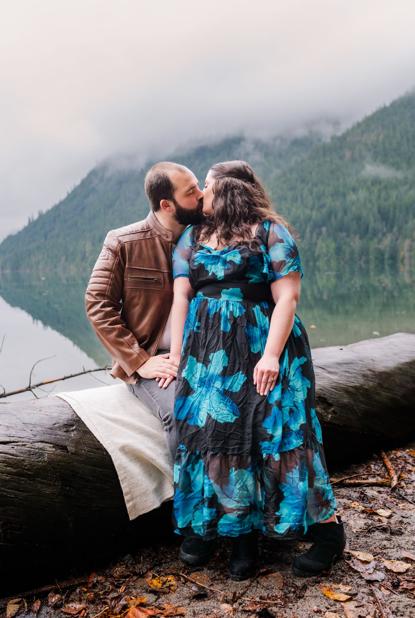Engaged couple kissing on a log by a misty mountain at Chilliwack Lake, woman in blue floral dress, man in brown leather jacket.