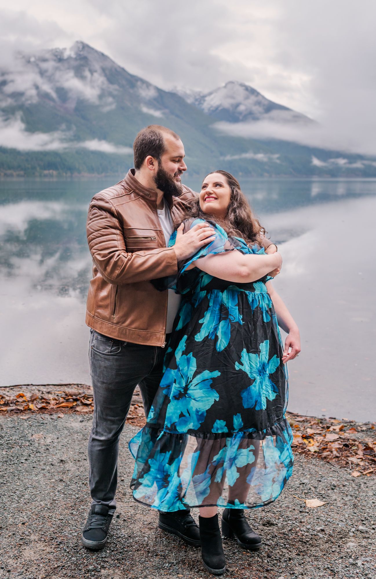 Engaged couple embracing by a reflective Chilliwack lake, woman in blue floral dress, man in brown leather jacket, misty peaks backdrop.