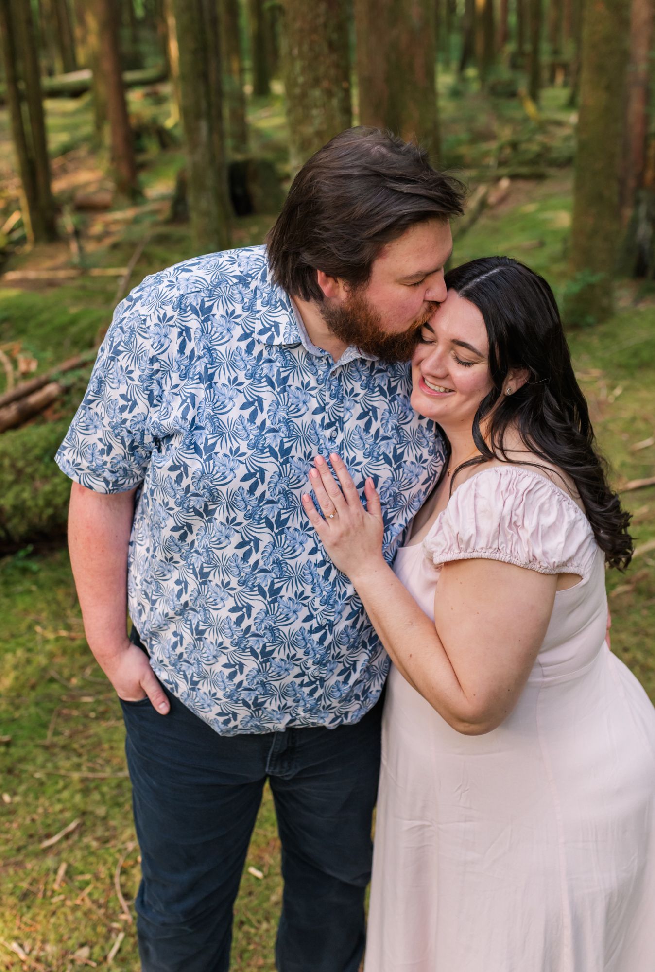 A Chillliwack engagement session shares a tender moment in a mossy forest, man in blue floral shirt kissing woman's head as she smiles.