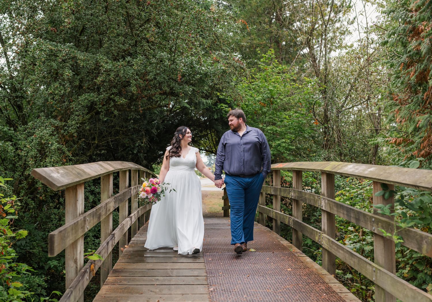 Bride and groom walking hand in hand on a wooden bridge surrounded by lush green trees on their Fraser Valley wedding day.