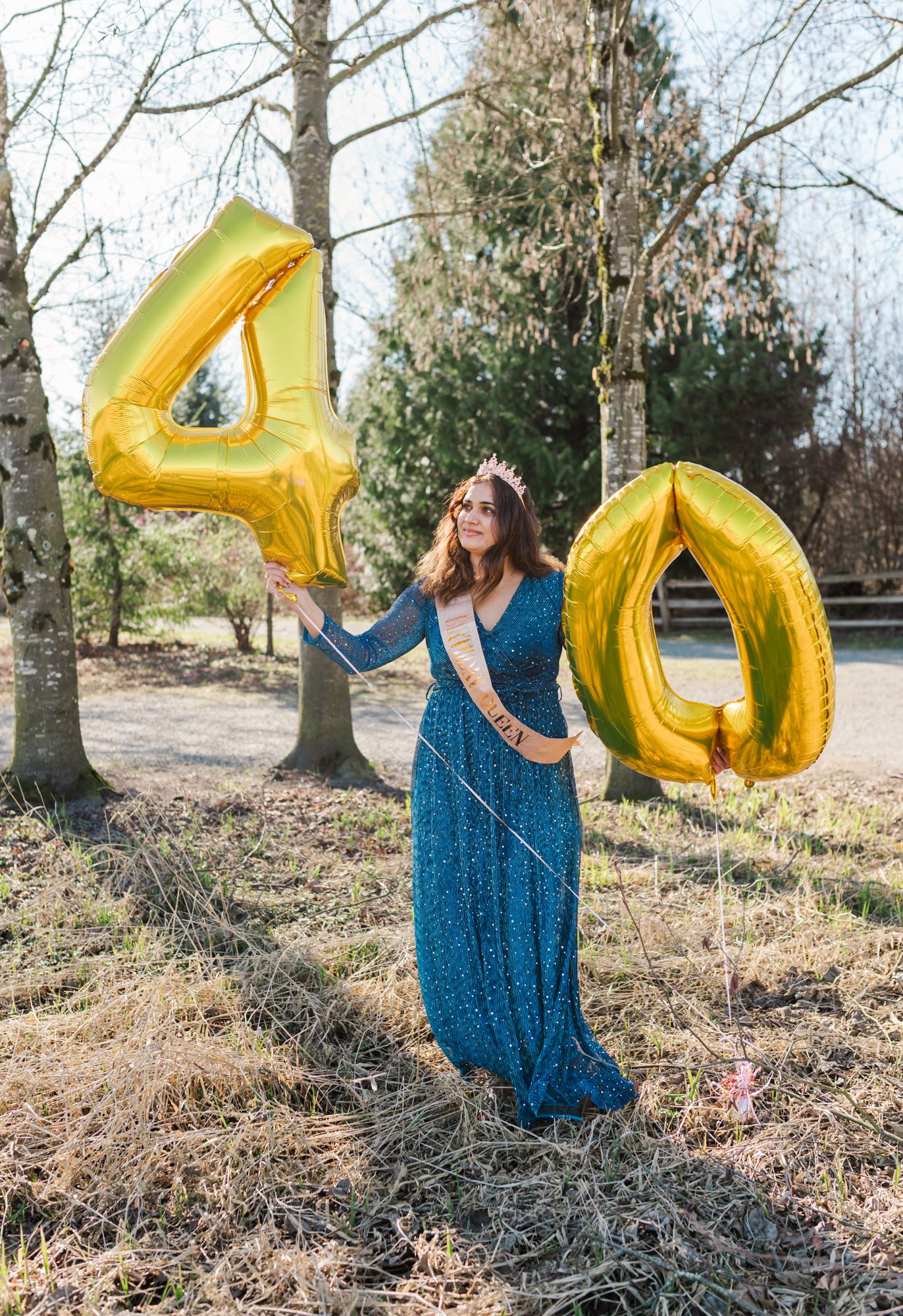 Woman in blue gown holding gold number 40 balloons outdoors, celebrating her 40th birthday with a tiara and sash for a Abbotsford session.