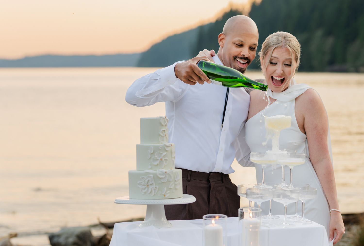 Bride and groom pouring champagne at outdoor wedding reception with tiered white cake and scenic waterfront sunset backdrop in Maple Ridge.
