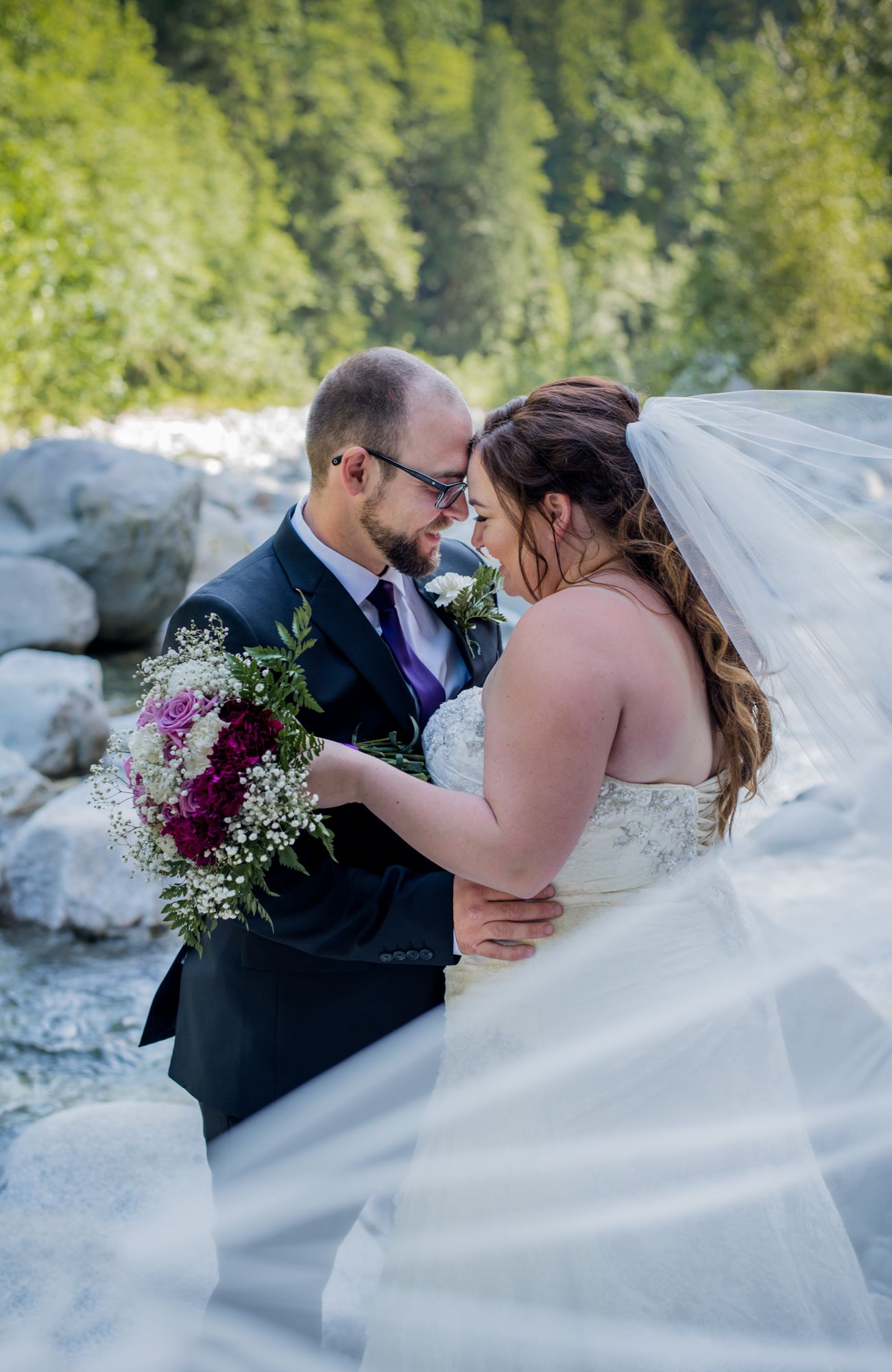 Bride and groom share romantic moment outdoors, bride holding purple bouquet with veil flowing, rocky forest backdrop in Mission, BC.
