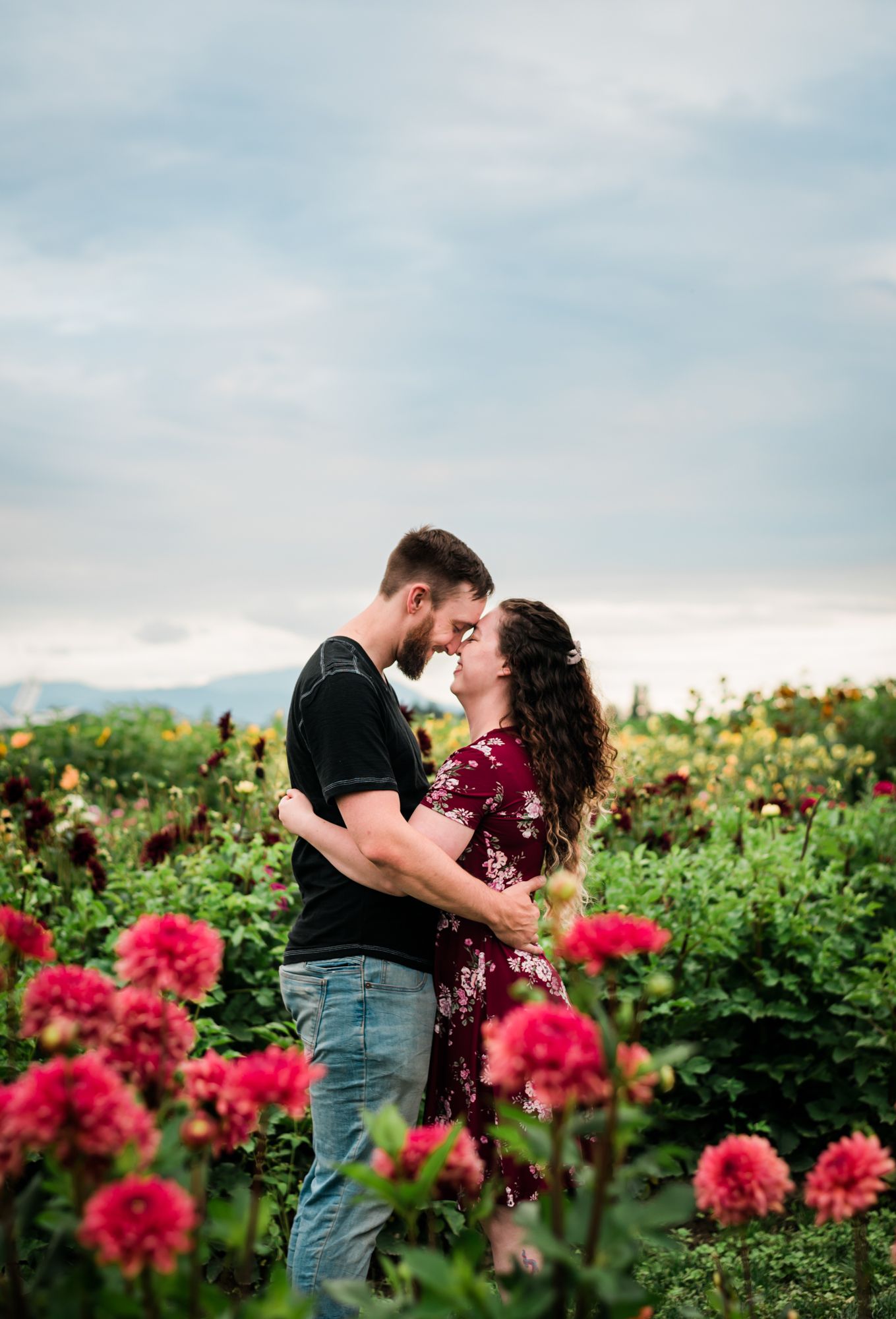 Couple embracing romantically in a vibrant flower garden with pink dahlias and lush greenery under a cloudy sky for a Chilliwack engagement.