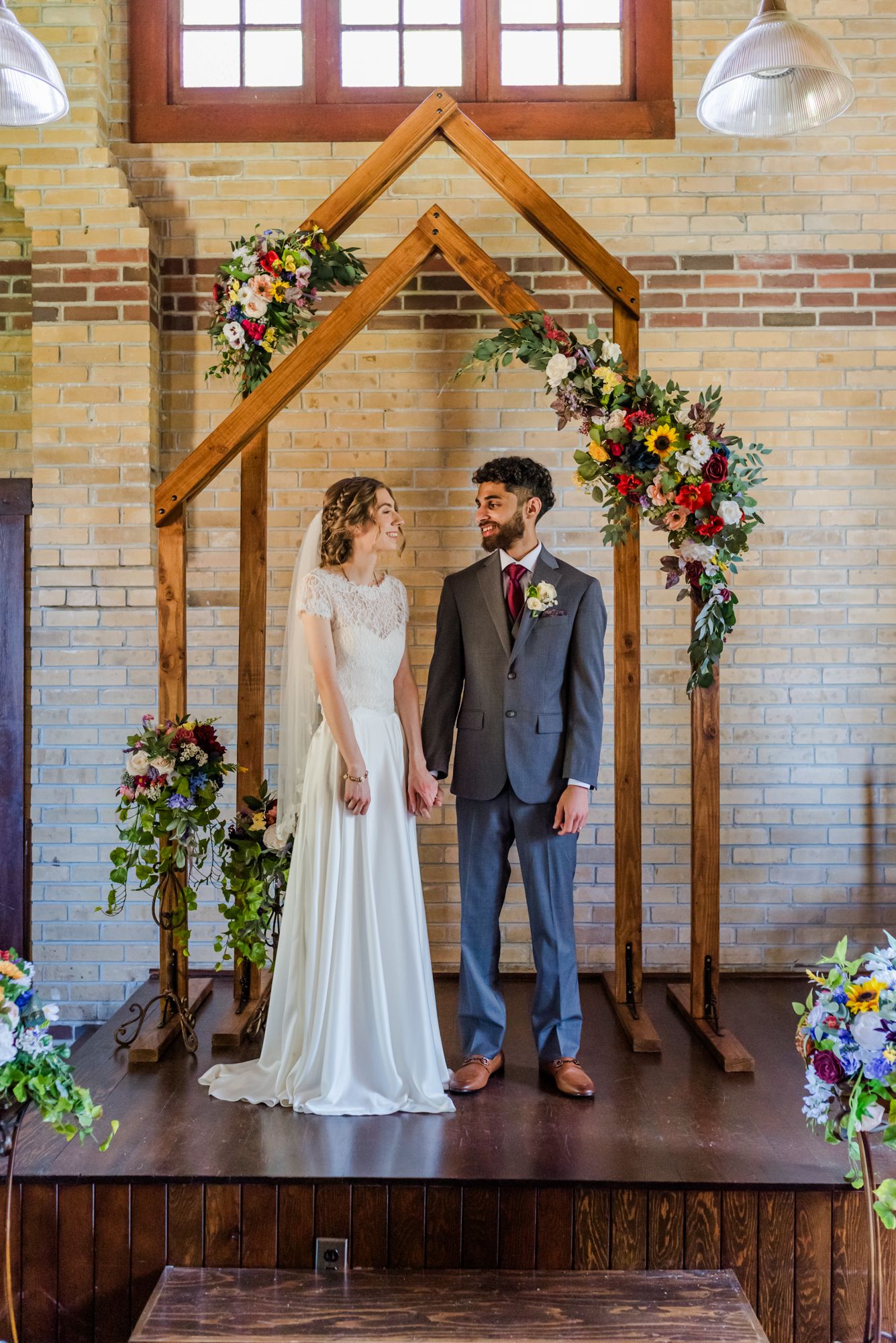 Bride and groom stand holding hands under a rustic wooden arch adorned with colorful flowers in a brick Abbotsford venue.