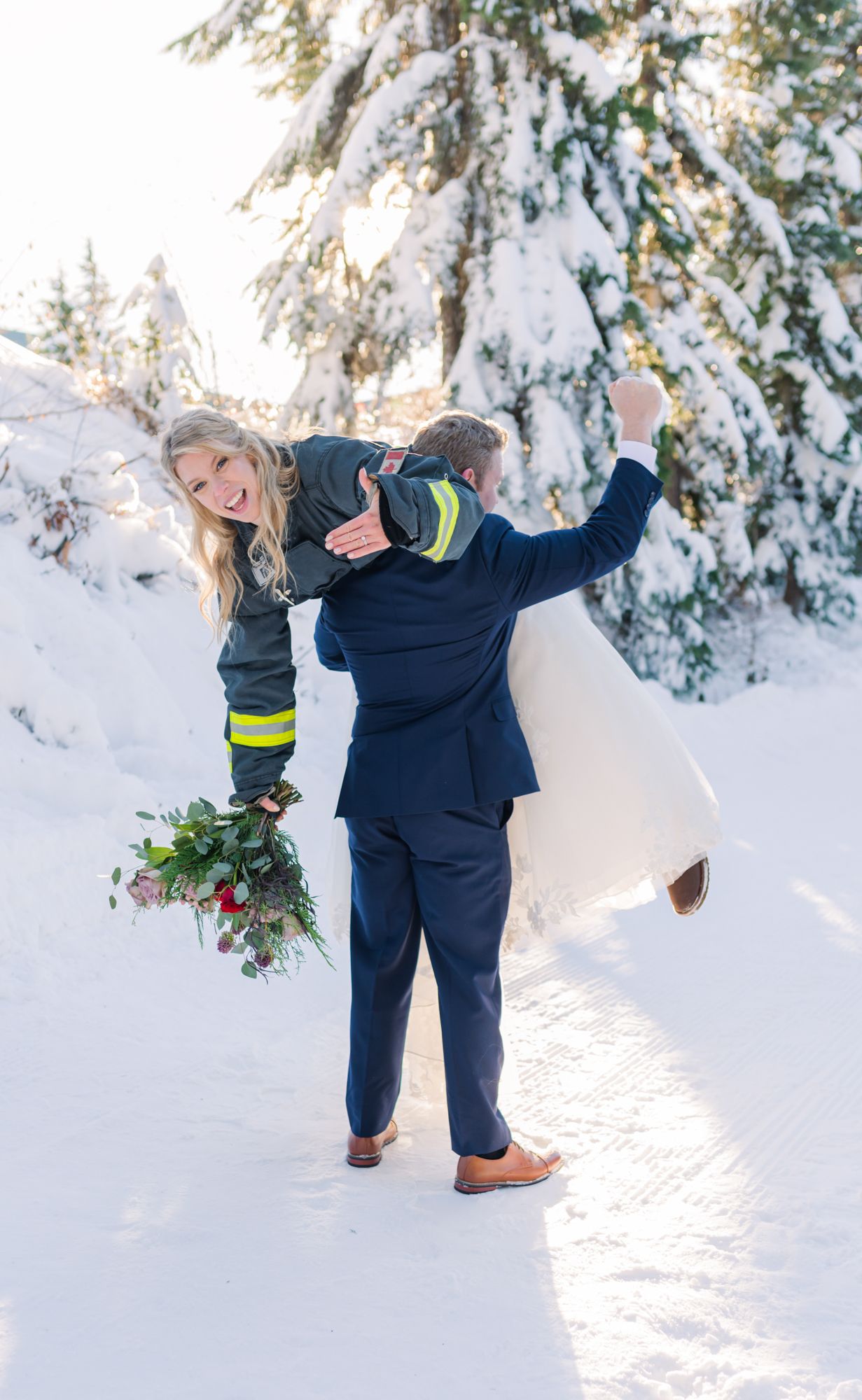Grouse Mountain elopement groom in navy suit carries bride in white dress over snowy path, pumping fist in celebration.