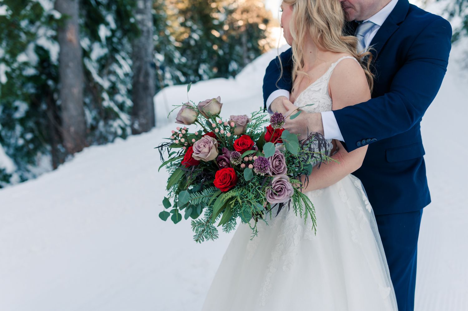 Wedding couple embracing in snowy forest, holding a lush red and pink rose bouquet with greenery in winter wedding attire at Grouse Mountain