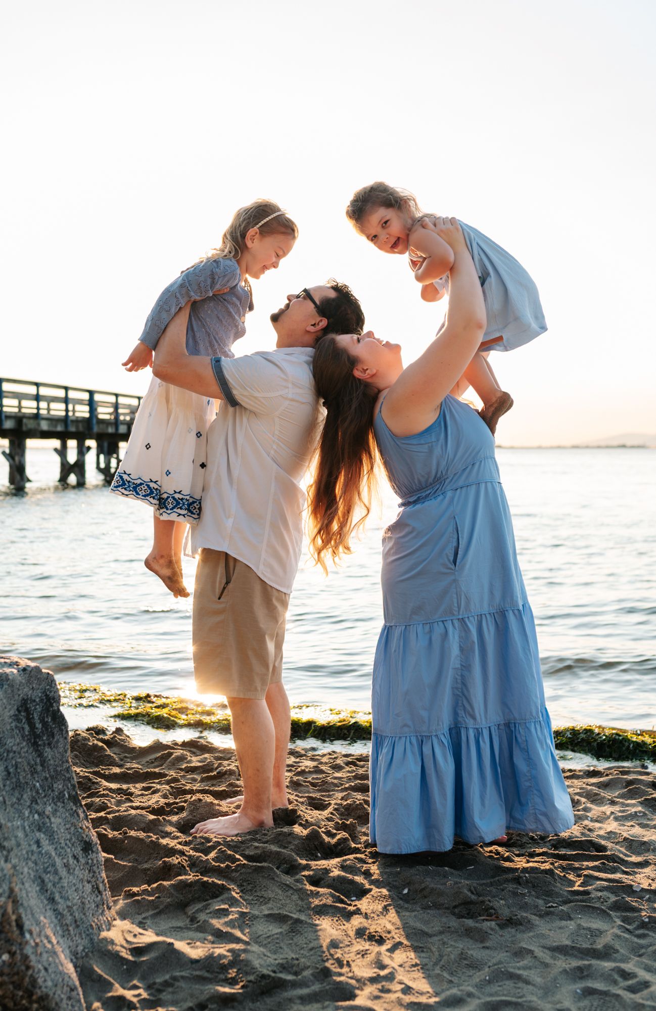 Family of four enjoying a sunny beach day, parents lifting two young children in matching blue dresses near a pier in Surrey BC.