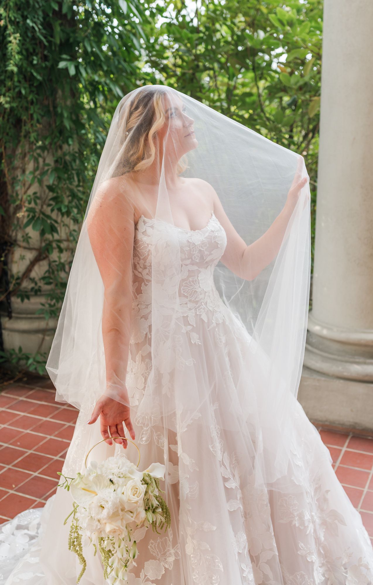 Bride in lace ball gown and flowing veil holds white bouquet outdoors near stone columns and greenery at Fraser Valley wedding.