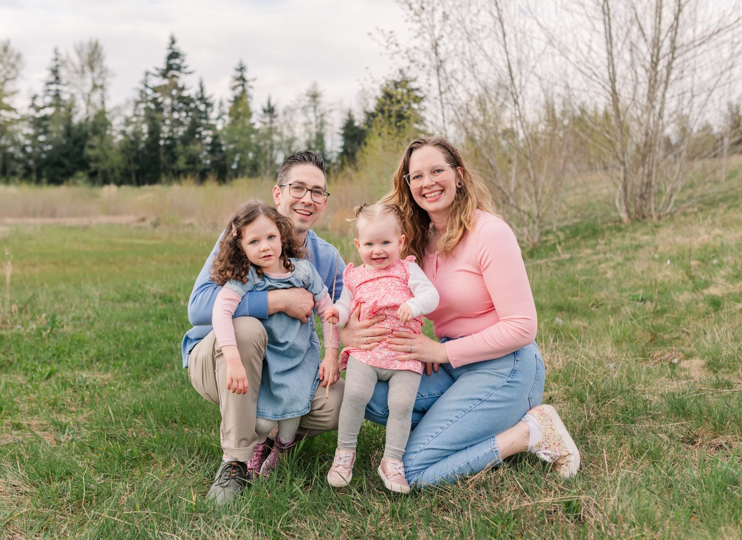 Happy family of four posing outdoors on green grass with trees in background during spring photo session in Abbotsford, BC.