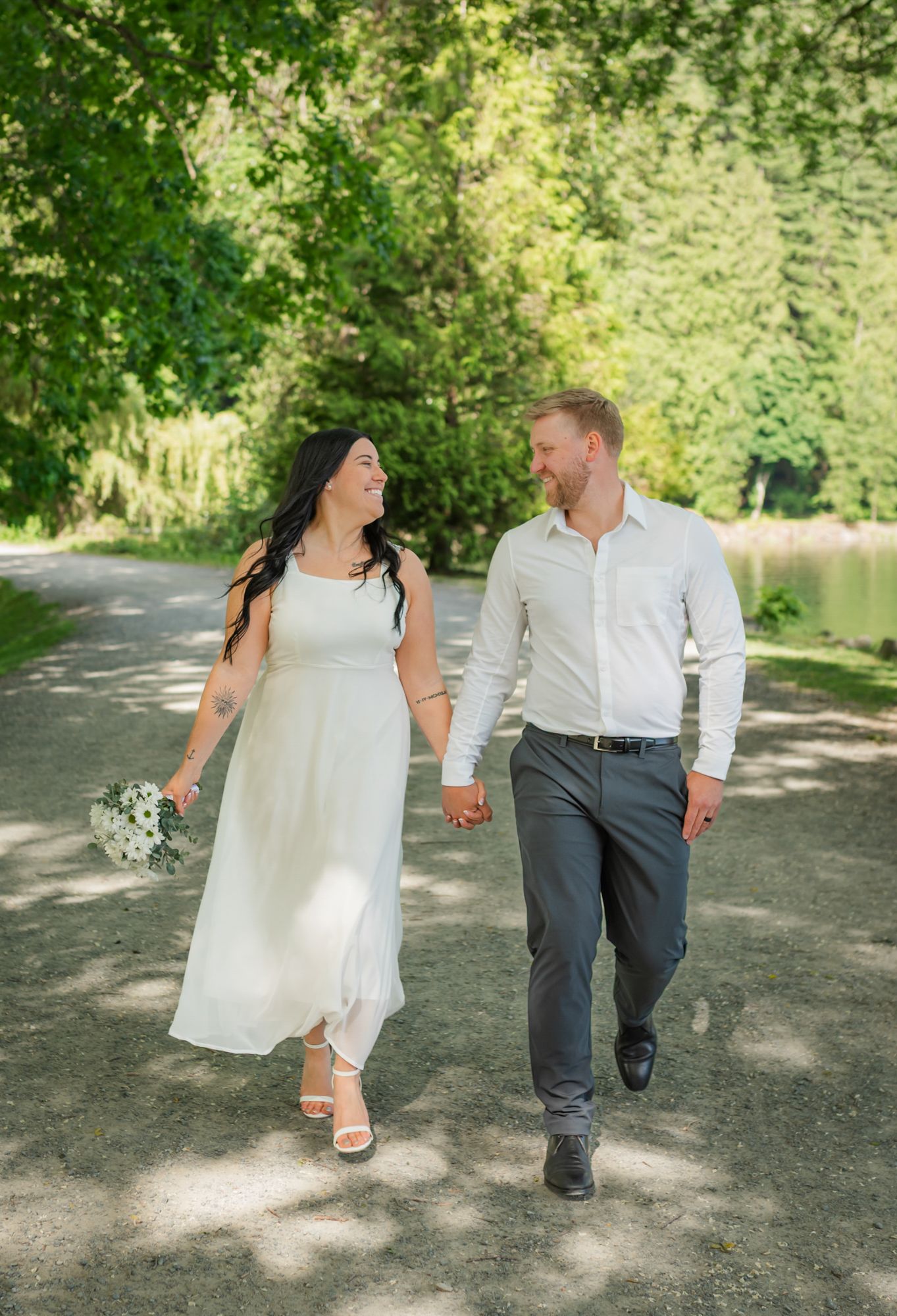 A smiling couple in wedding attire walks hand-in-hand along a sunlit park path surrounded by lush green trees in Harrison Hot Springs, BC.