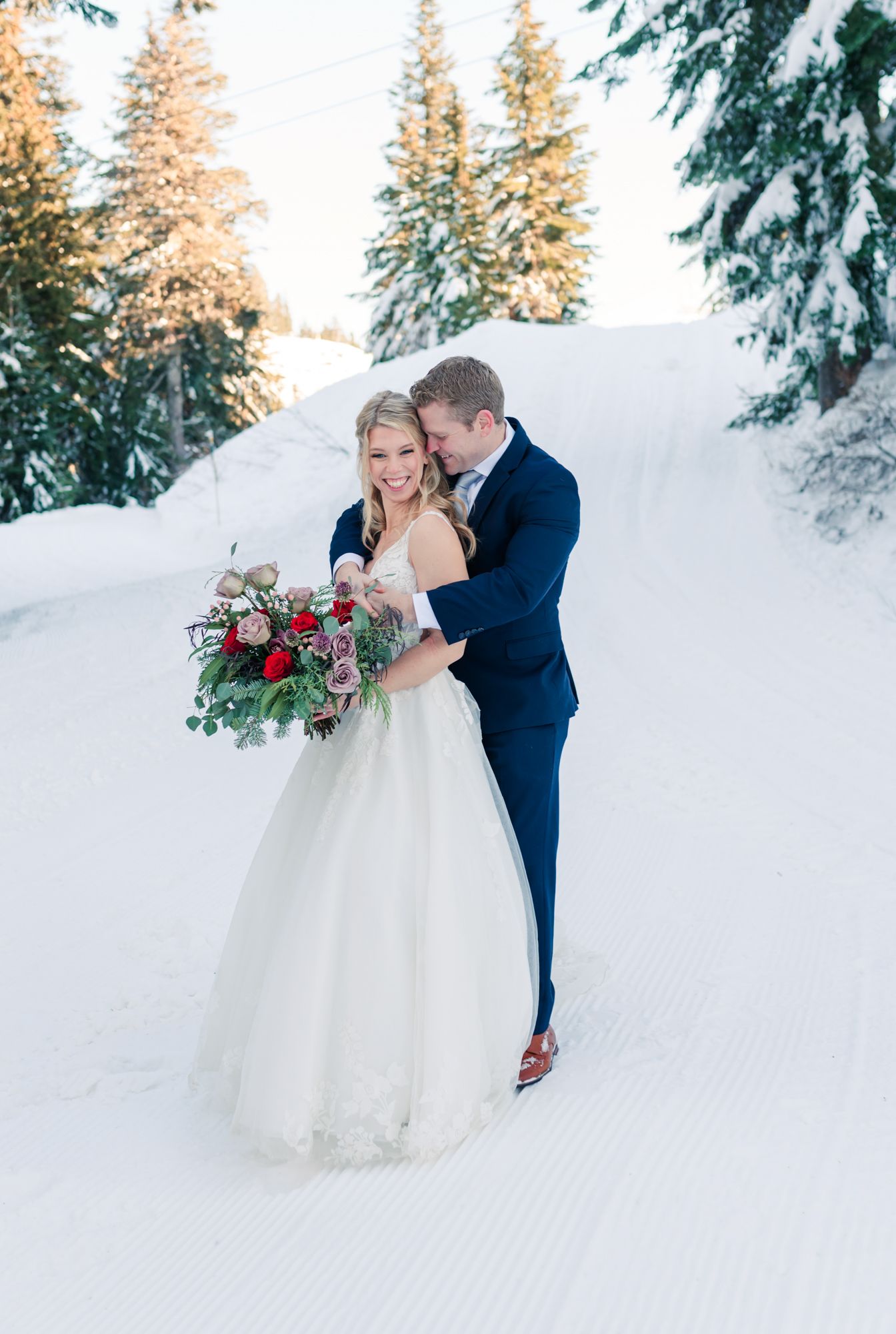 Just married couple in snowy winter wonderland elopement, she holds red floral bouquet, he wears navy suit, trees behind at Grouse Mountain.