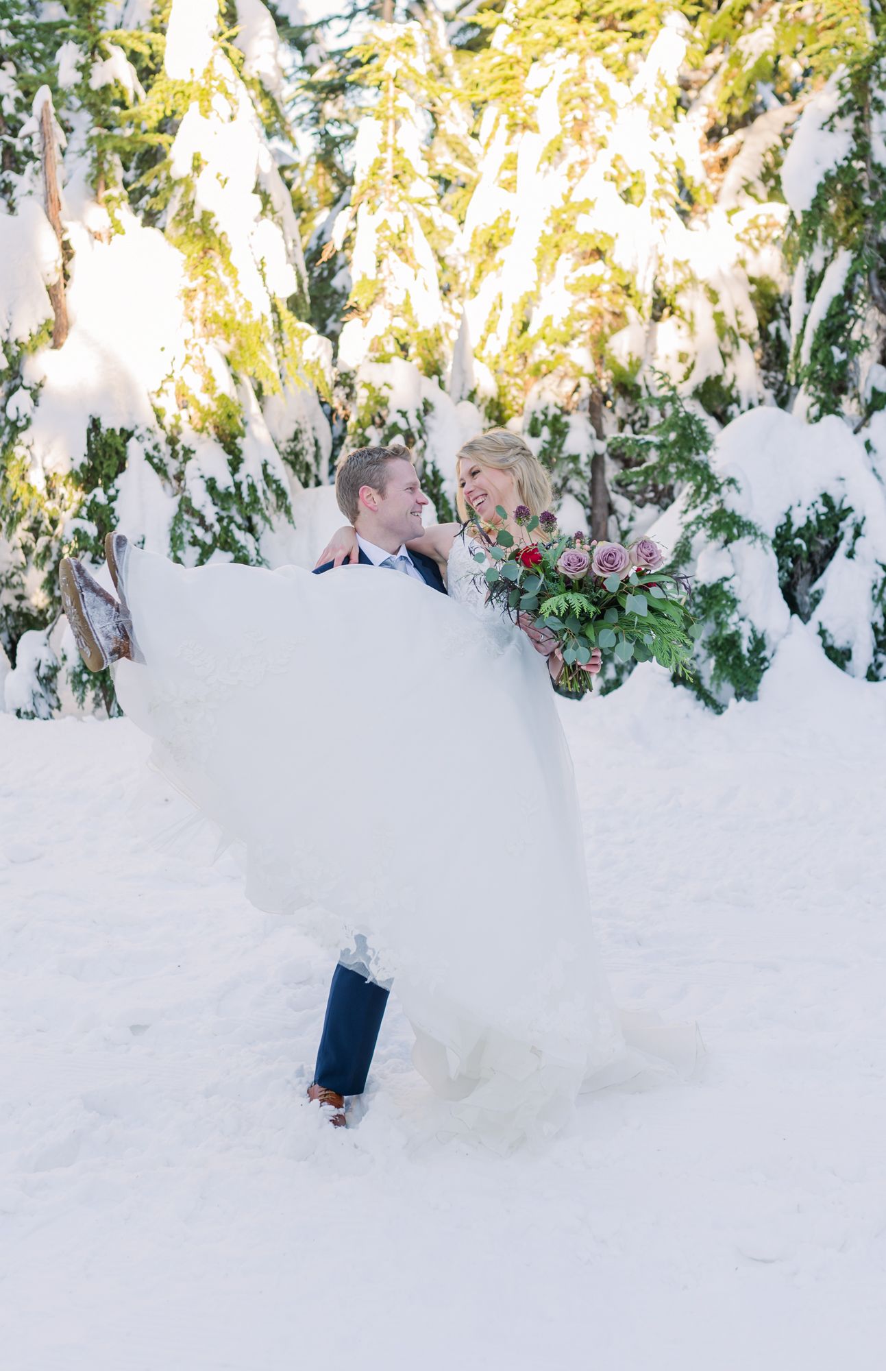 Groom lifting bride in white gown amid snow-covered evergreen trees in a romantic winter wedding outdoor portrait at Grouse Mountain.