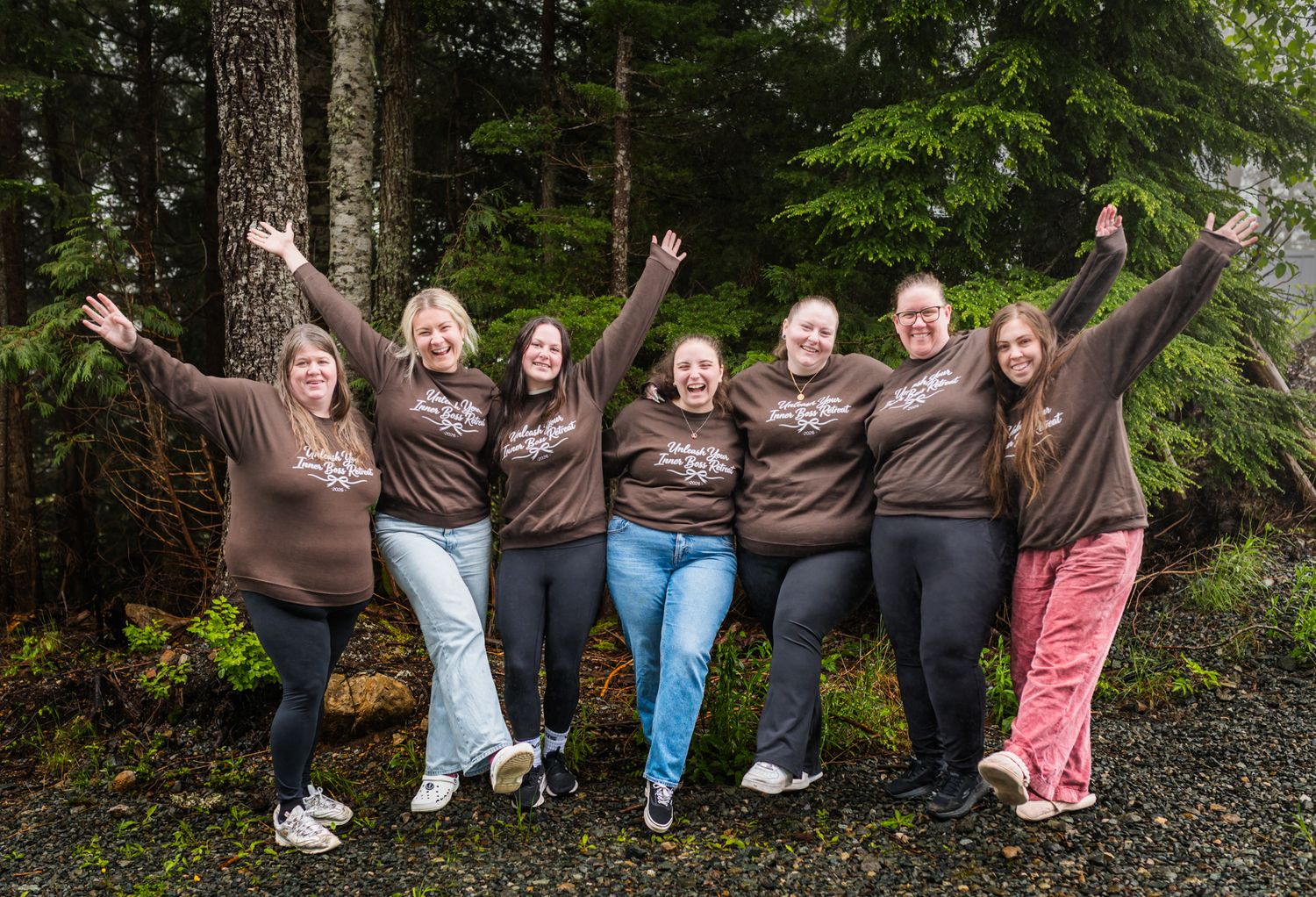 Seven women in matching brown shirts pose cheerfully outdoors among evergreen trees, arms raised in celebration retreat in Agassiz, BC.
