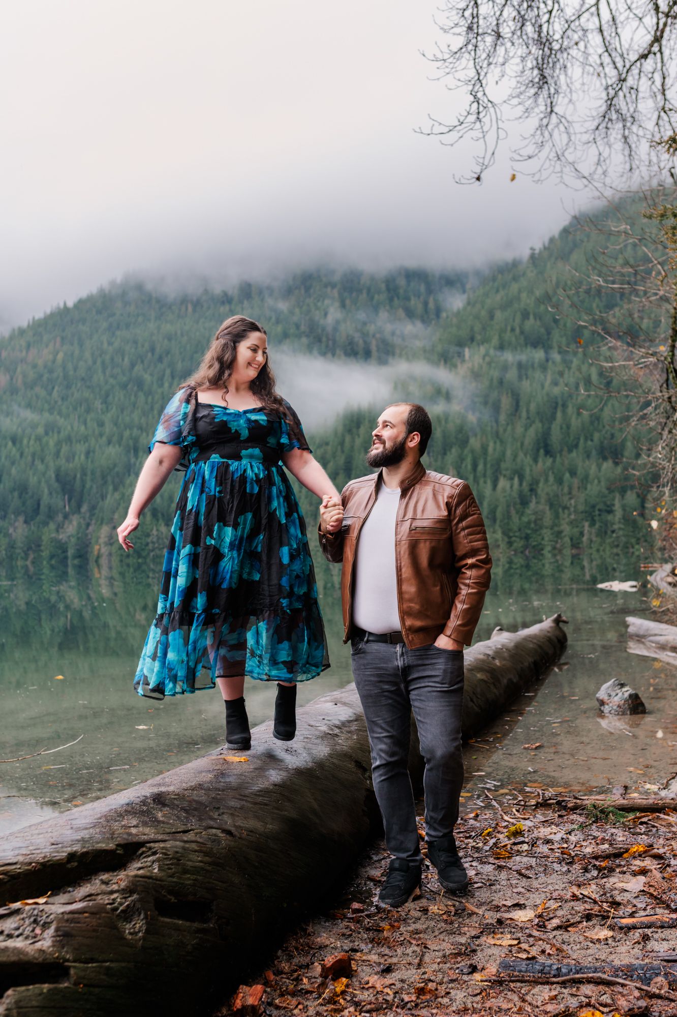 Couple holding hands on a log by a misty mountain at Chilliwack lake, woman in blue floral dress, man in brown leather jacket.