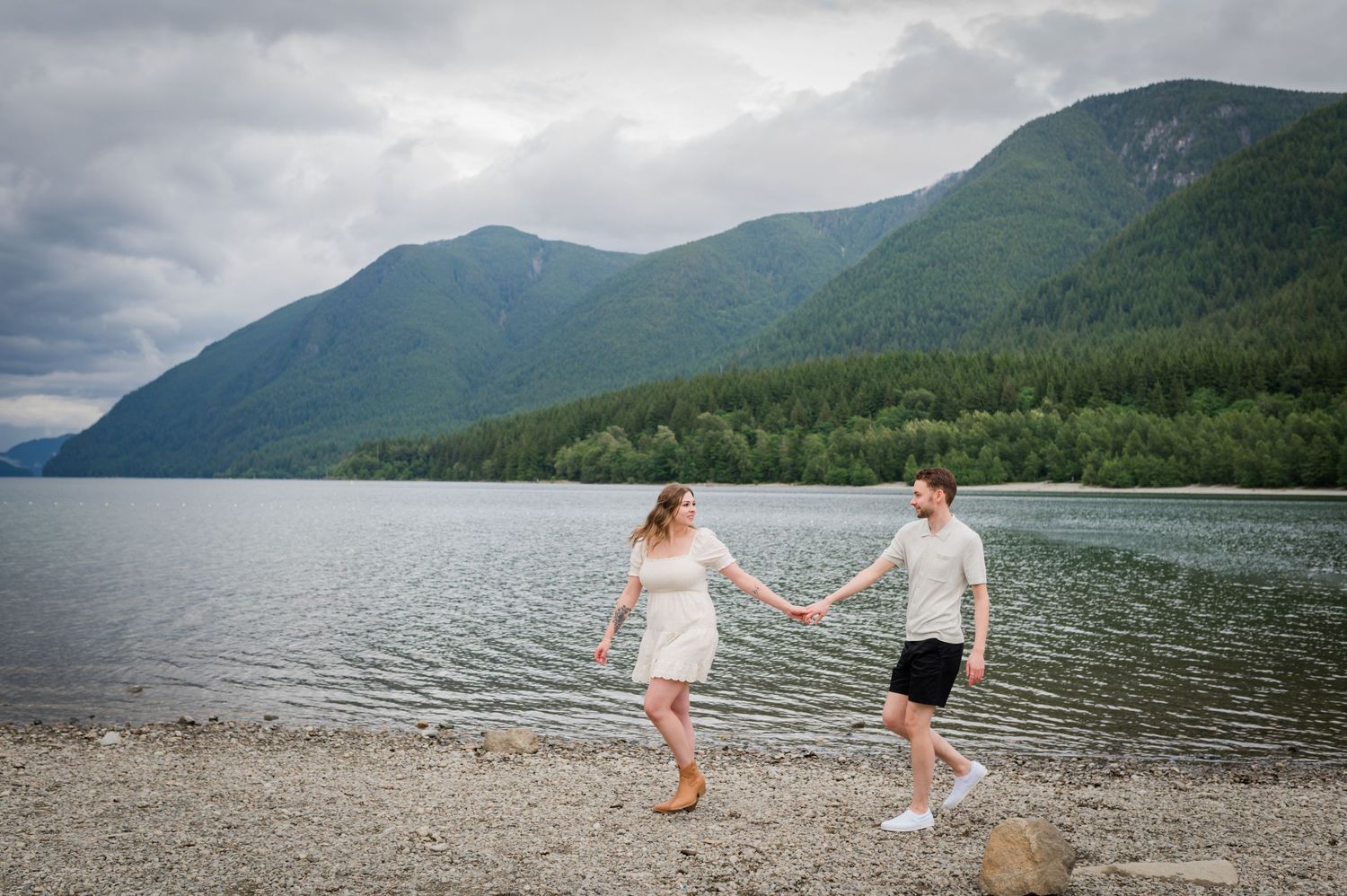 Couple holding hands walking along a rocky lakeside beach with lush green mountains and cloudy skies in the background at Alouette Lake.