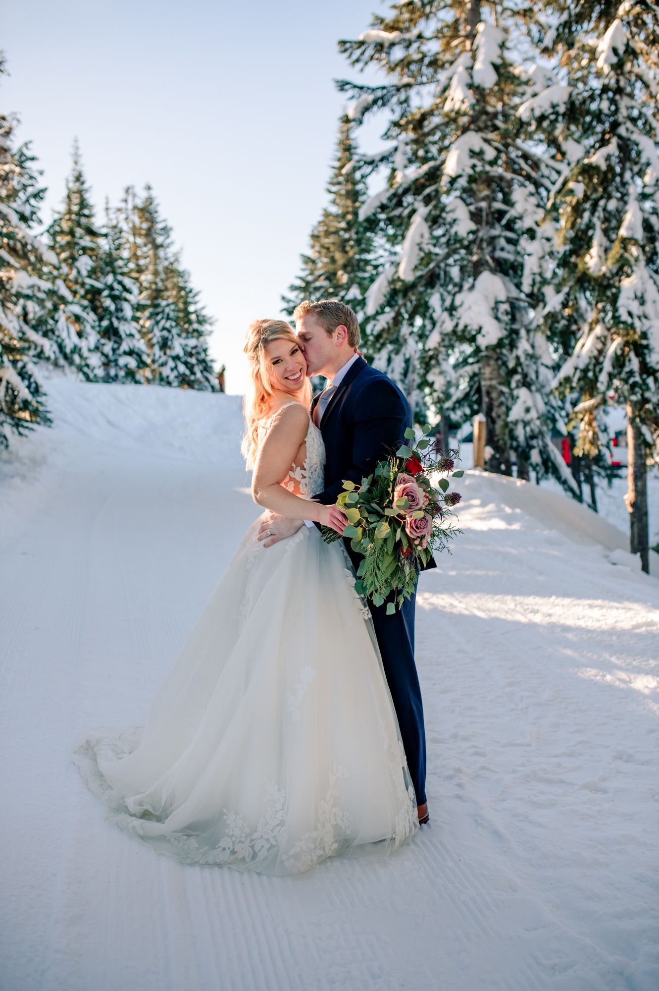 Grouse mountain wedding bride and groom embrace in snowy winter wonderland setting with snow-covered pine trees and bridal bouquet.