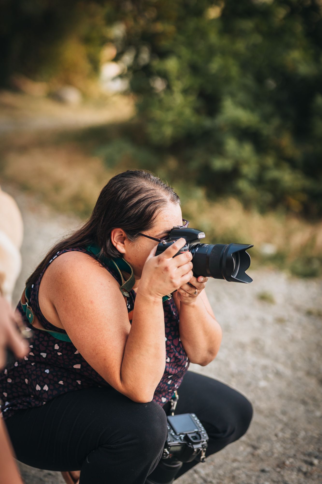 Fraser Valley photographer crouching outdoors, holding a camera up to her eye, photographing in a natural setting with trees in background.