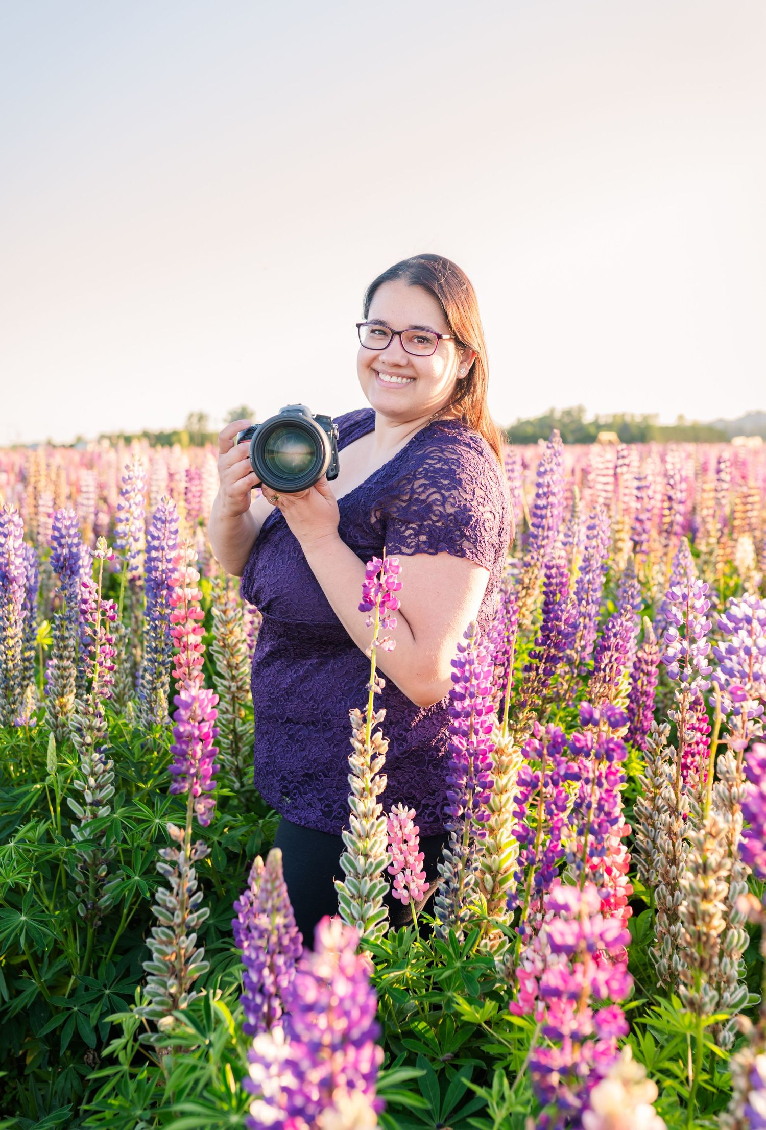 Fraser Valley photographer smiling in a vibrant lupine flower field holding a DSLR camera, wearing glasses and a purple lace top.