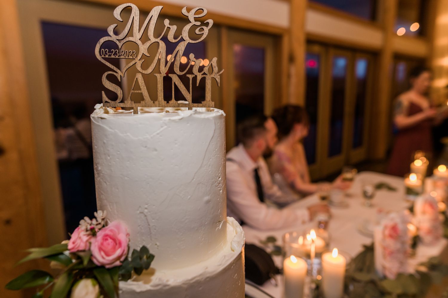 White two-tier wedding cake with Mr & Mrs Saint topper, pink roses, and candles at elegant reception.