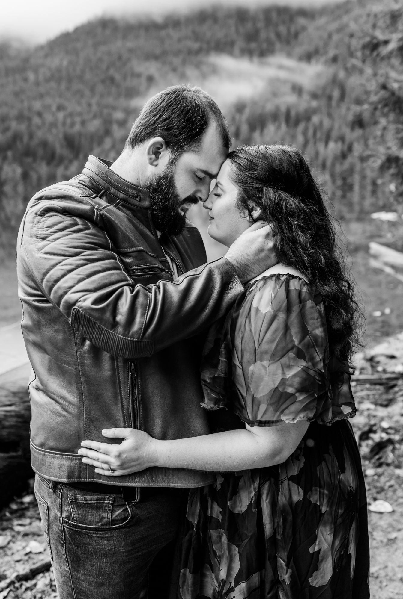 Black and white photo of a bearded man and curly-haired woman embracing tenderly outdoors with misty mountains behind them Chilliwack Lake.
