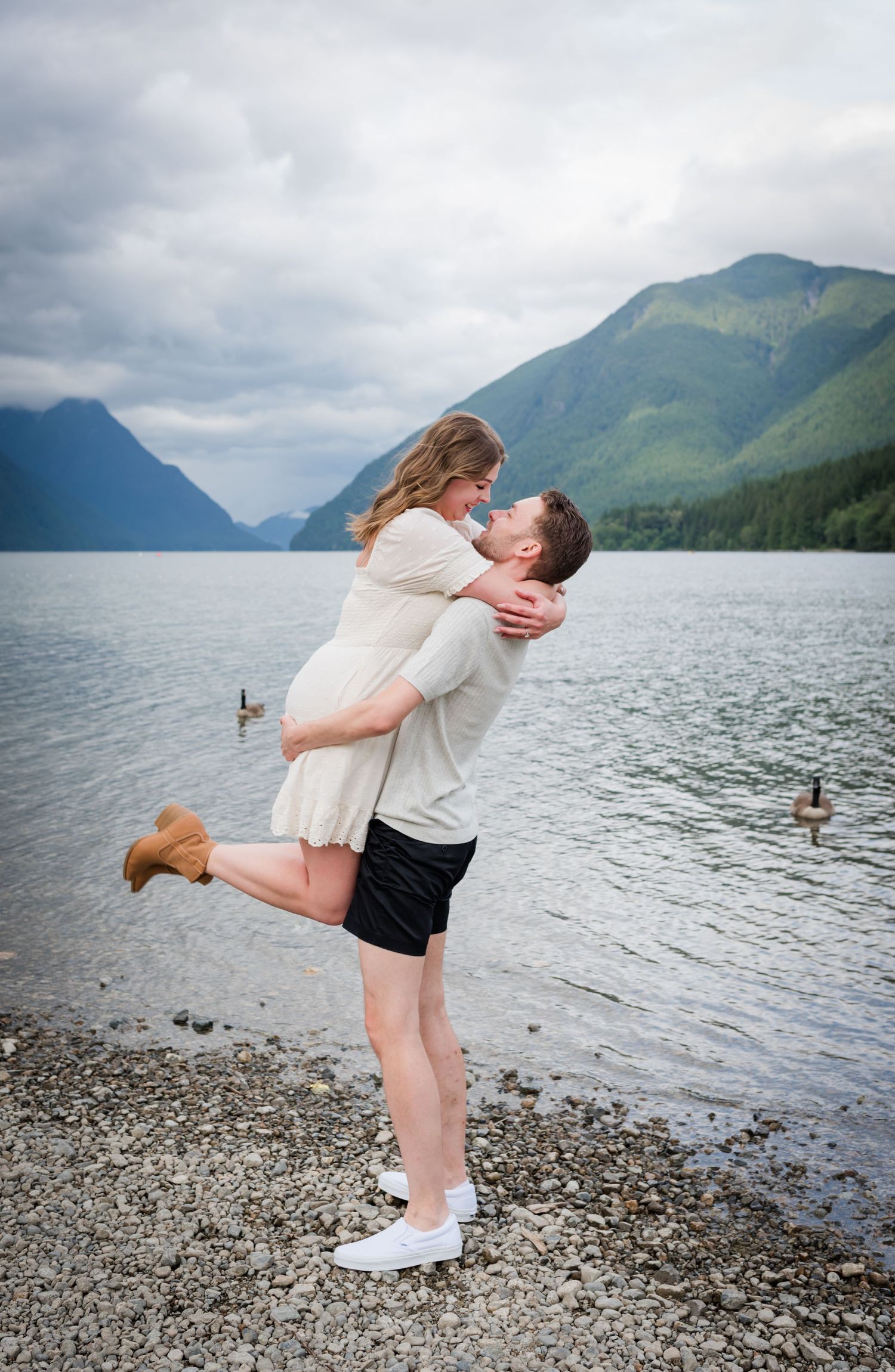 A man lifts a woman in a romantic embrace on a rocky lakeshore with misty mountains in the background.