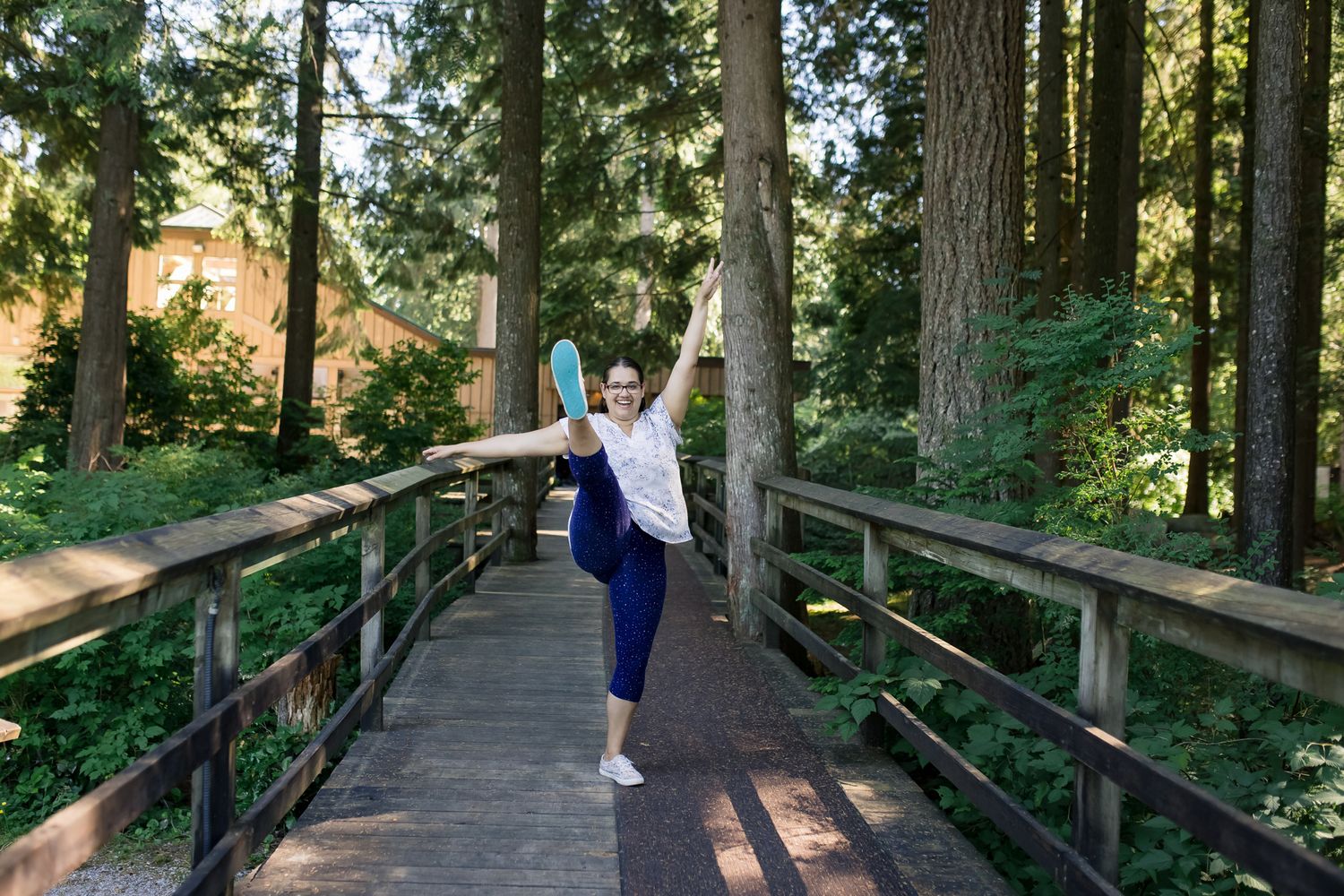 Fraser Valley photographer in blue pants balancing on one leg on a wooden bridge surrounded by lush green forest trees.