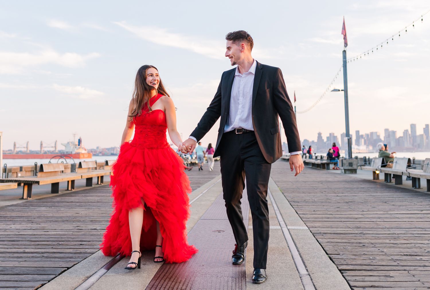 Couple walking on a waterfront pier, woman in red ruffled gown and man in black suit, city skyline in background for a Vancouver engagement.