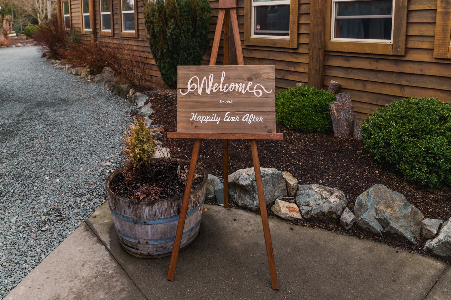 Wooden welcome sign on easel reading Happily Ever After at outdoor wedding venue entrance with barrel planter at Fraser River Lodge.